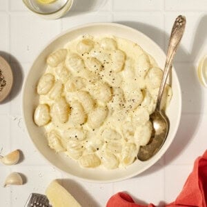 a cream colored plate with creamy sauce with gnocchi and a spoon. The bowl is surrounded by garlic clove, parmesan cheese, a microplane, red napkin, and glasses