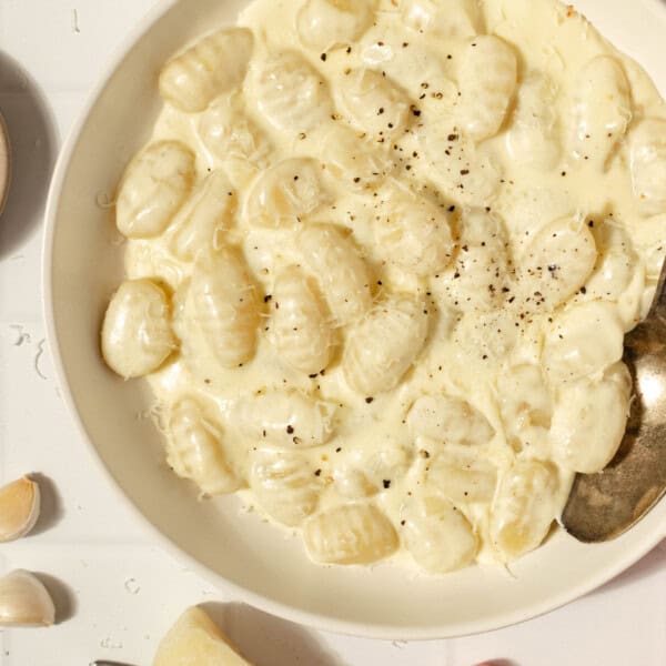 overhead photo of a bowl of gnocchi in a creamy sauce with a spoon. a red towel and ingredients surround the bowl