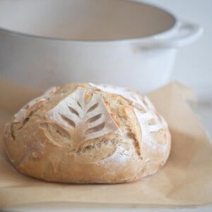 A sourdough loaf on parchment paper with a white dutch oven in the background.