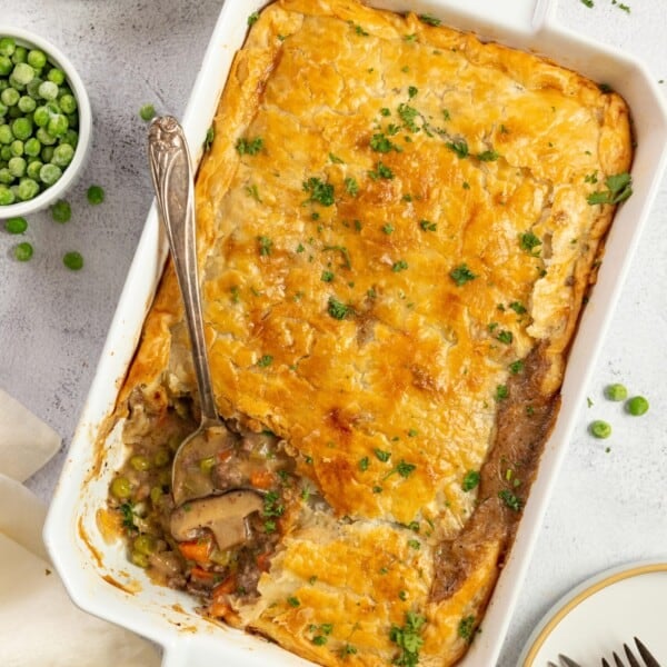 overhead photo of ground beef pot pie topped with a golden brown pie crust and parsley with a spoon in the corner of the pie. The baking dish sits on a white countertop surrounded by ingredients