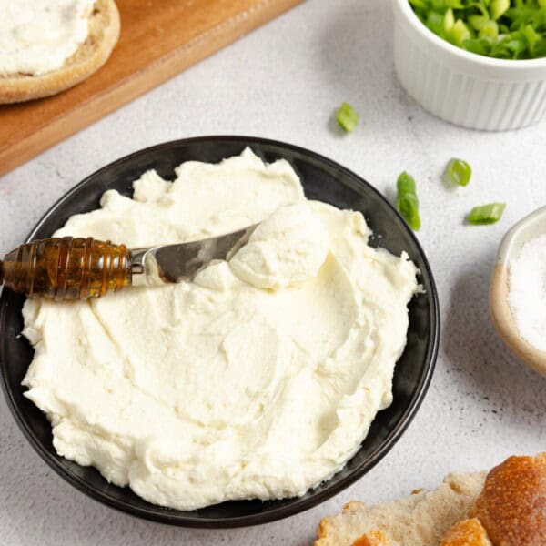 a bowl of homemade cream cheese with a spreading knife in it and other food to eat with the cream cheese surrounding it on a white countertop and wooden cutting board