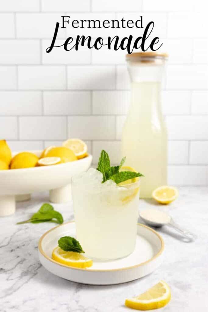 glass of fermented lemonade topped with mint leaves on a white plate. A bowl of lemons and a tall jar of lemonade are in the background