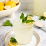 glass of lemonade topped with a slice of lemon and mint leaves on a white plate with lemons, mint, and a tall jar of fermented lemonade in the background