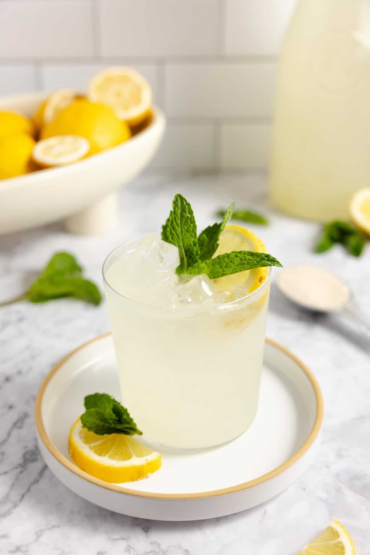 glass of lemonade topped with a slice of lemon and mint leaves on a white plate with lemons, mint, and a tall jar of fermented lemonade in the background