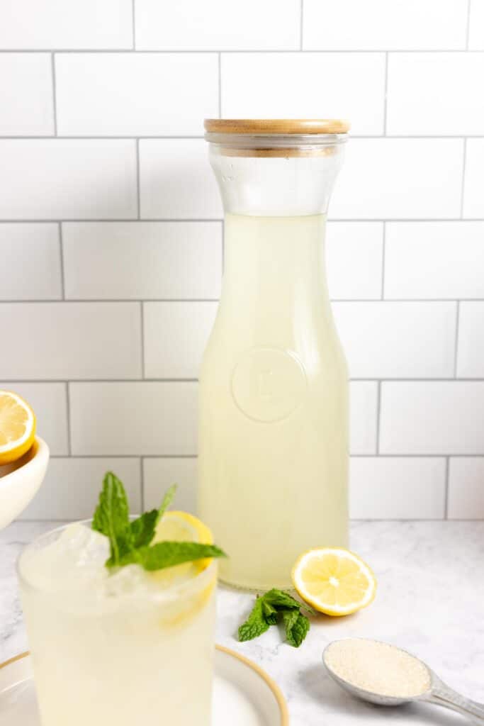 a cup and a large jar of fermented lemonade on a white countertop