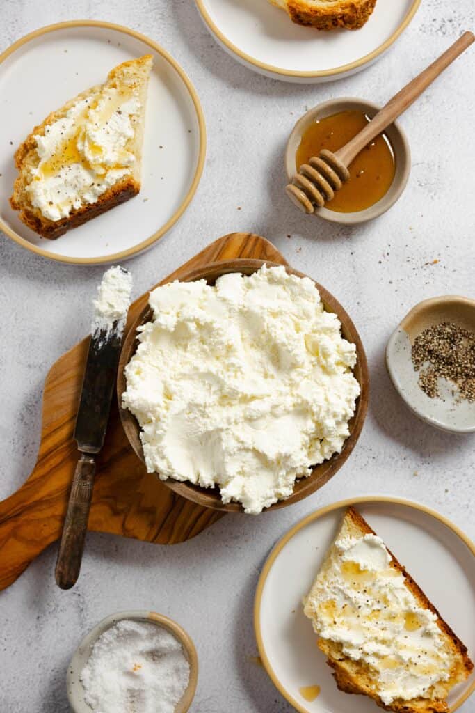 wooden bowl full of ricotta cheese with small plates full of honey, toast, salt, and more surrounding the bowl