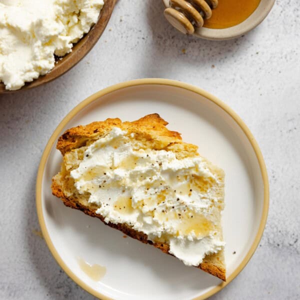 ricotta cheese slathered on toast and topped with honey on a white plate with honey and more ricotta in the background