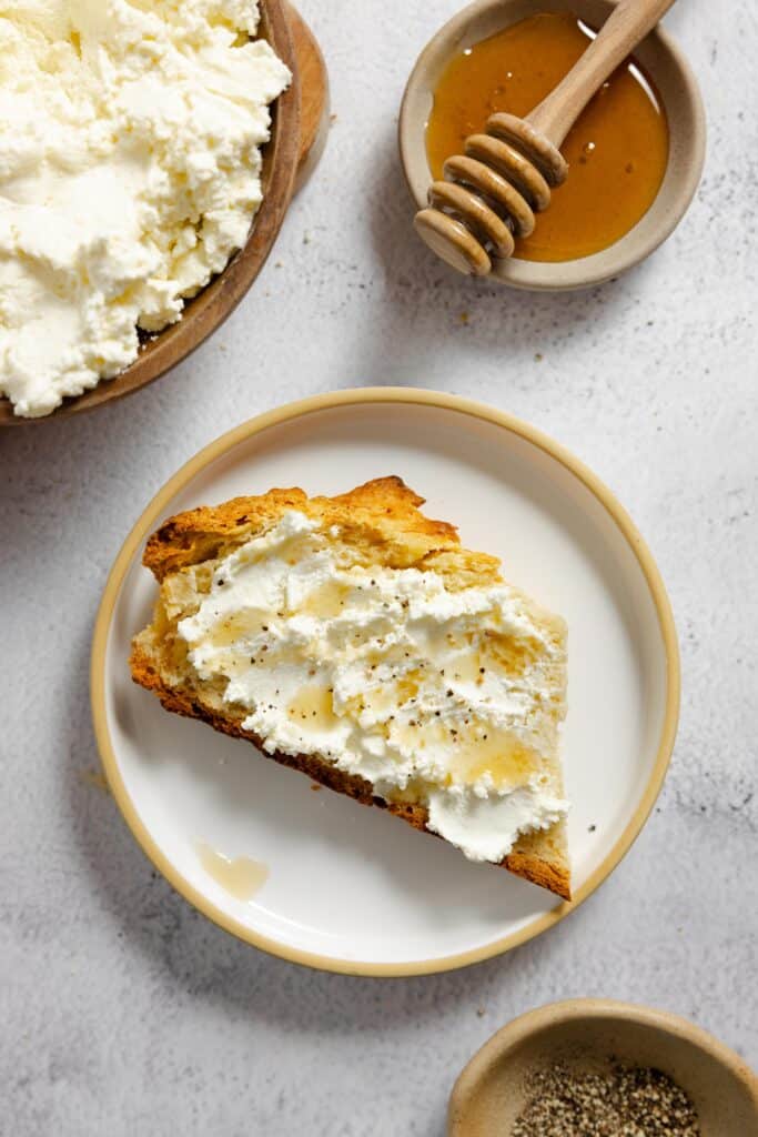 ricotta cheese slathered on toast and topped with honey on a white plate with honey and more ricotta in the background 