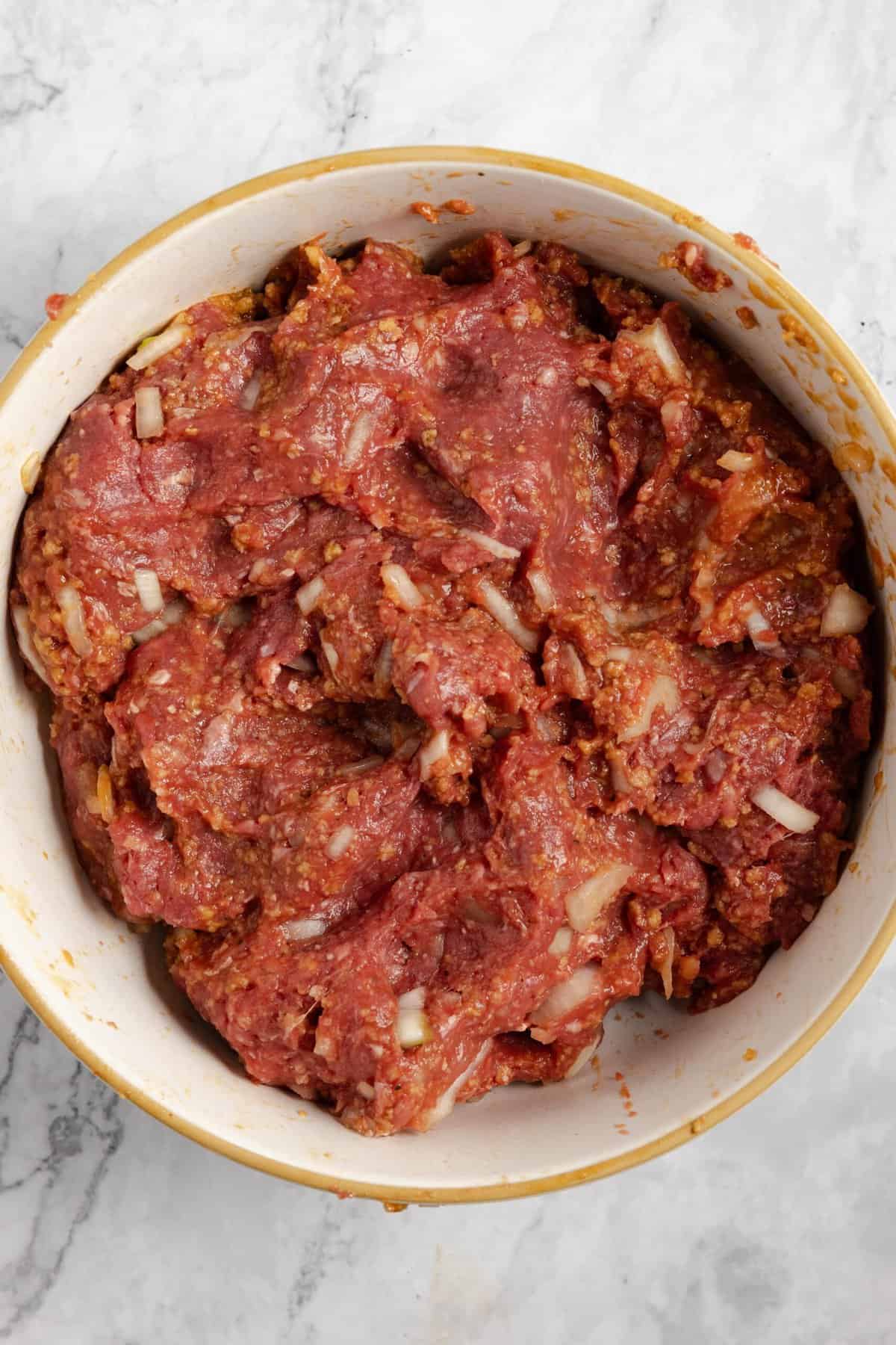 All of the ingredients for meatloaf mixed together, including ground beef, in a large bowl sitting on a white countertop