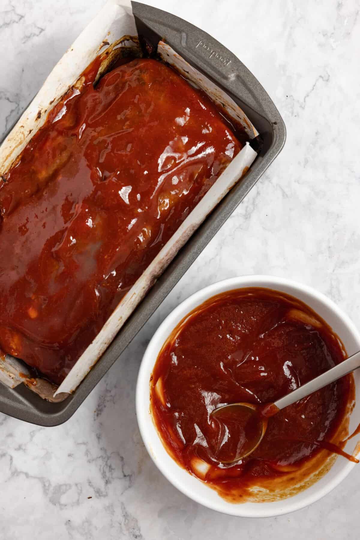 Meatloaf in a loaf pan with glaze on top with the bowl of glaze sitting next to it on a white countertop