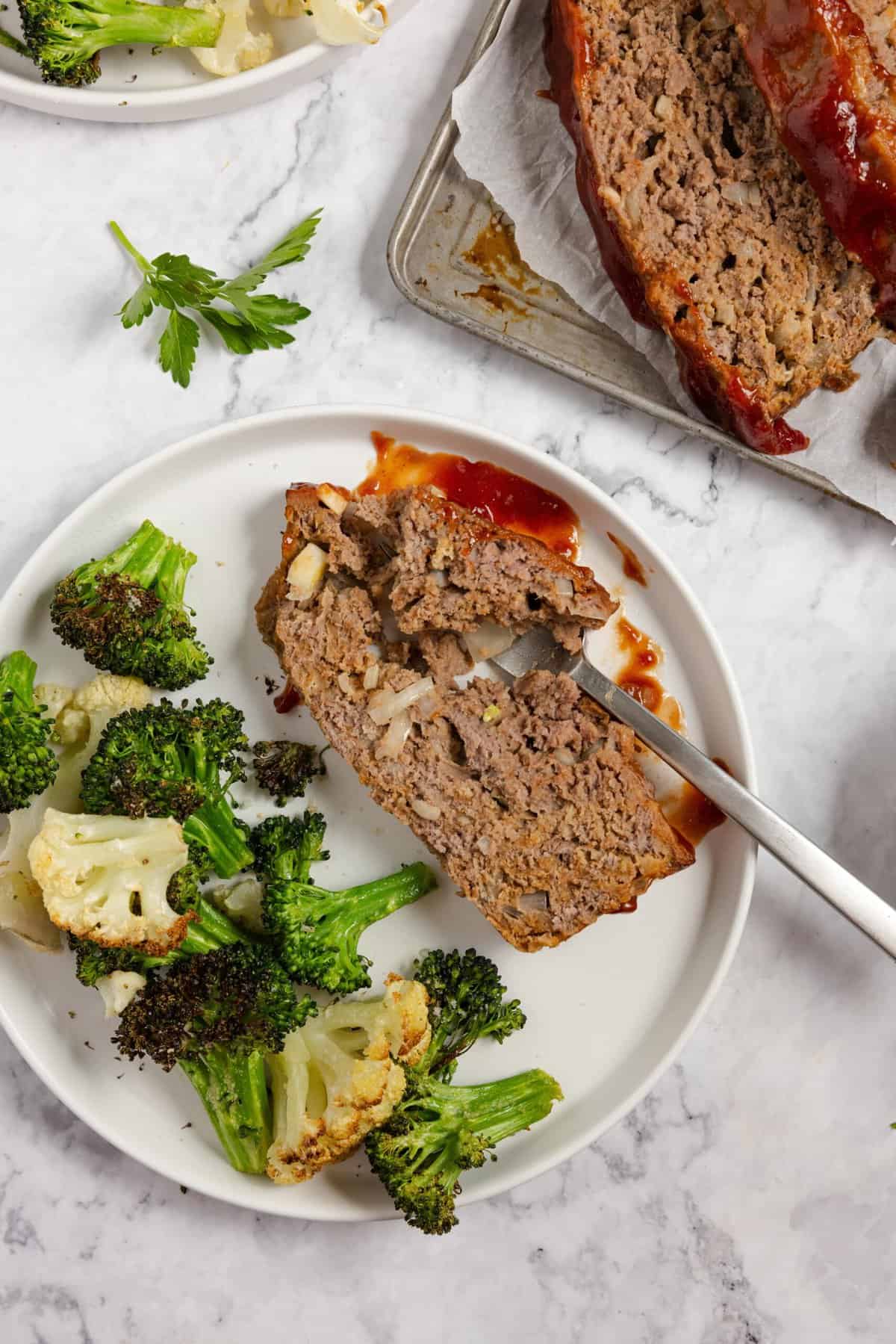 A plate with a slice of meatloaf with a fork slicing into it with a side of broccoli and cauliflower 