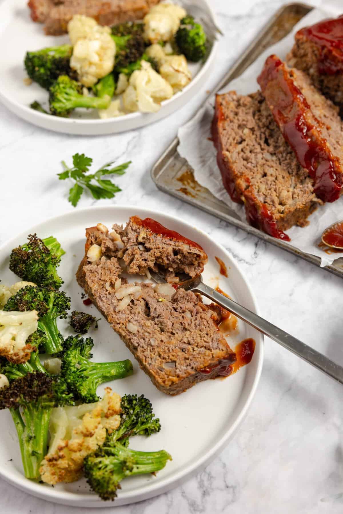 A plate with a slice of meatloaf and veggies on the side next to a platter with the rest of the meatloaf