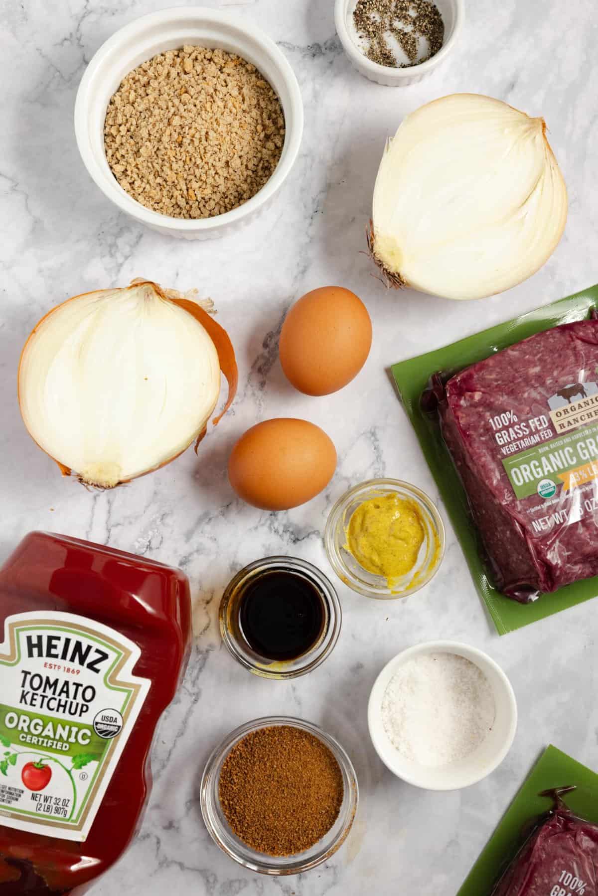 The ingredients for meatloaf spread out on a white countertop, including two eggs, ketchup, ground beef, onion, and spices. 