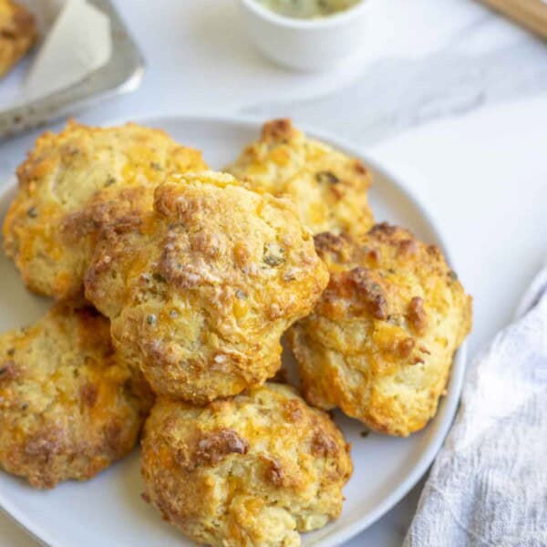 A white plate full of sourdough cheddar biscuits on a white countertop next to a tea towel and white bowl full of melted buttery and a pastry brush