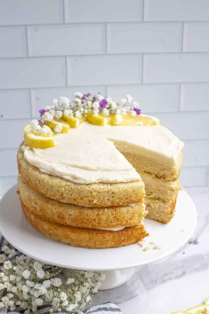 Sourdough lemon cake with a slice missing on a white cake platter with a white tile background