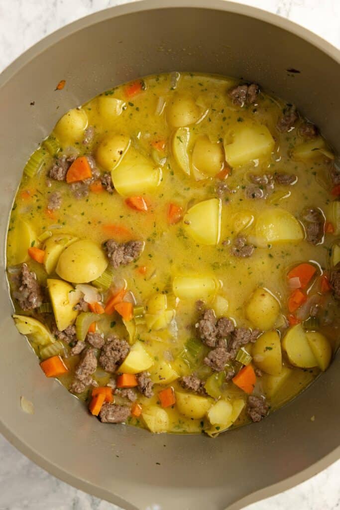 A soup pot filled with potatoes, ground beef, carrots, and bone broth simmering on the stove.