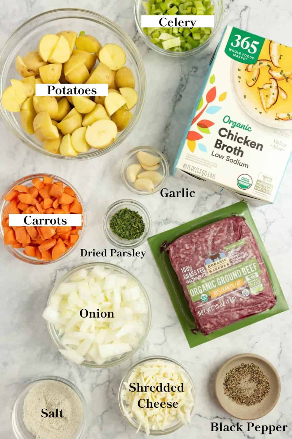 A box of chicken broth, a package of ground beef, bows filled with carrots, potatoes, onions, parsley and garlic on a white countertop.