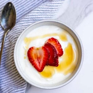 Raw milk yogurt topped with maple syrup and sliced strawberries in a bowl on a white and blue striped towel