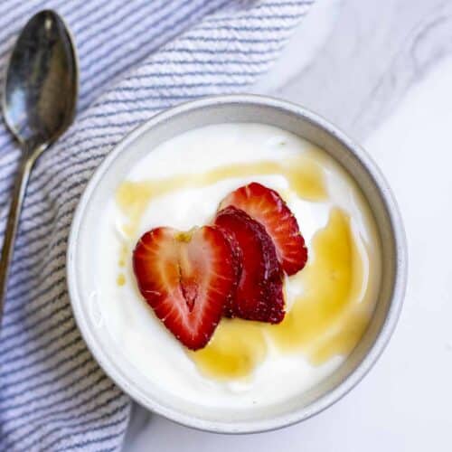 Raw milk yogurt topped with maple syrup and sliced strawberries in a bowl on a white and blue striped towel
