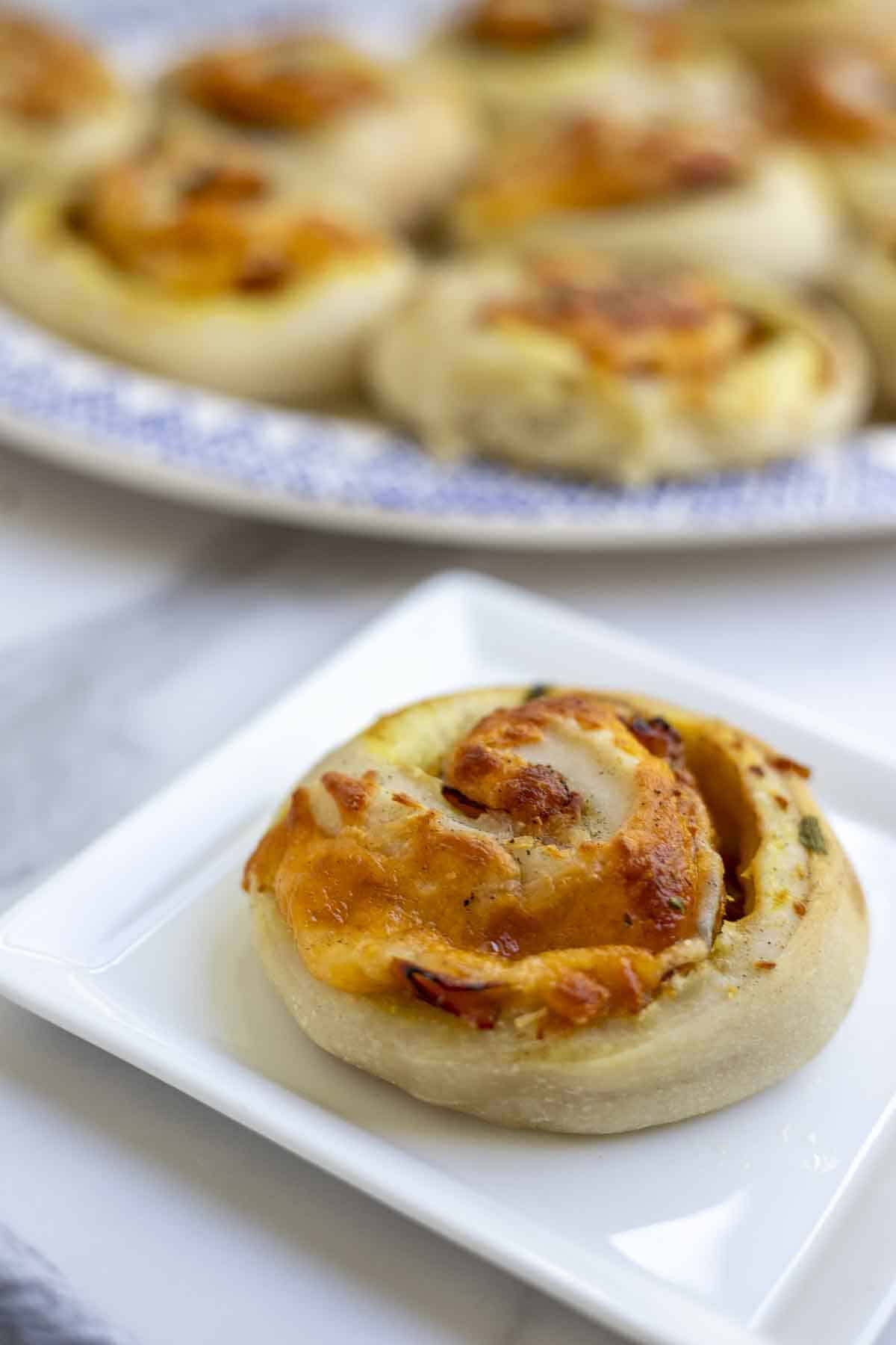 A sourdough ham and cheese roll on a white square plate with a platter of rolls in the background on a white marble countertop