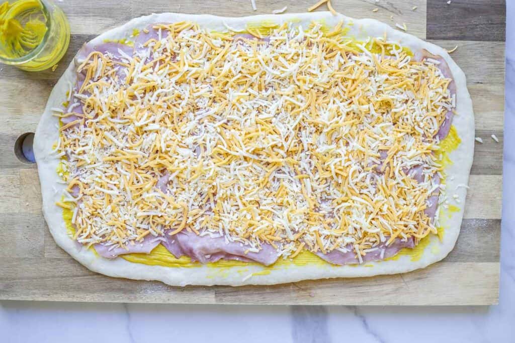 Sourdough ham and cheese roll dough rolled out on a wooden cutting board and brushed with mustard and layered with ham slices and shredded cheese