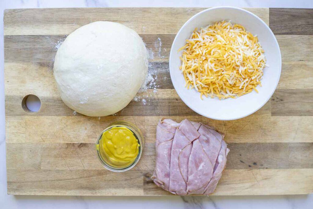 sourdough ham and cheese roll ingredients on a wooden cutting board, including shredded cheese in a bowl, sliced ham, mustard in a small bowl, and a ball of dinner roll dough