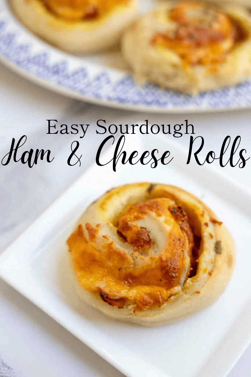 A sourdough ham and cheese roll on a square white plate on a white countertop with a platter of ham and cheese rolls in the background