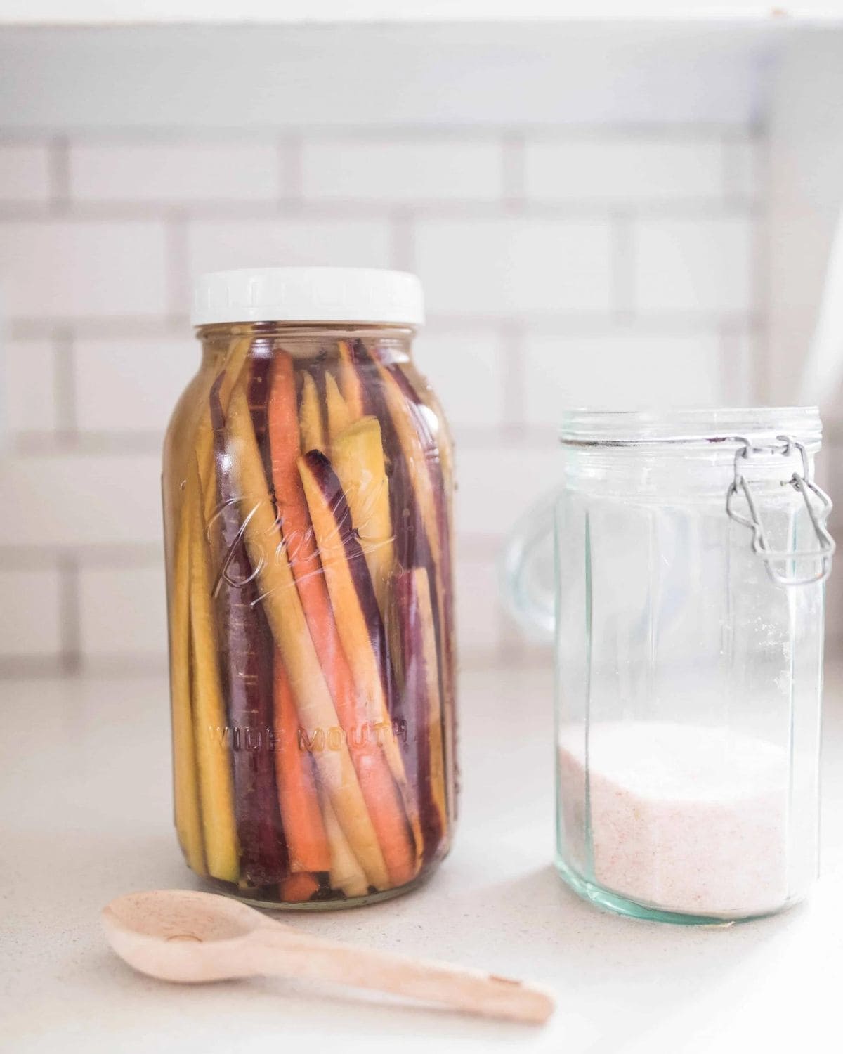 Large mason jar filled with tri-colored carrot sticks next to a jar of salt and a wooden spoon.