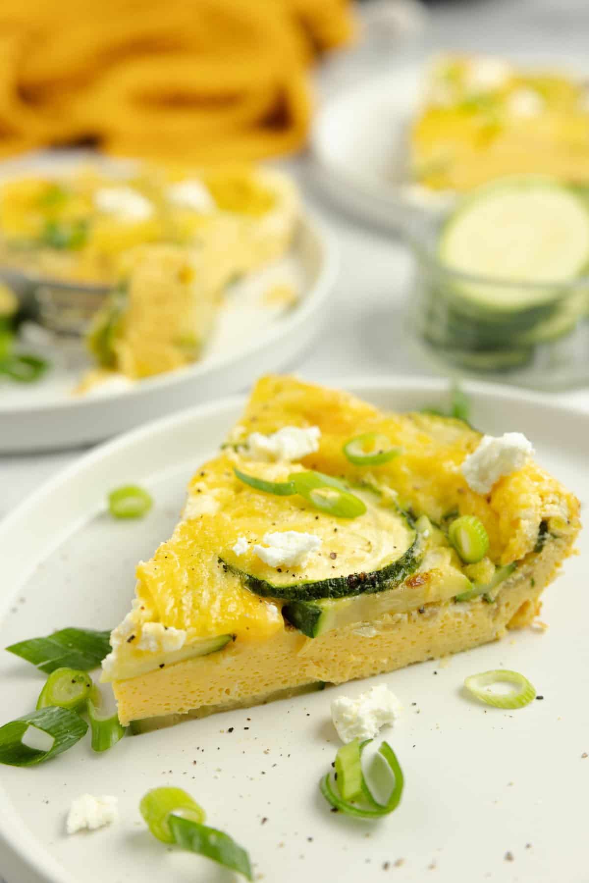 slice of zucchini frittata topped with herbs on a white plate with more frittata slices in the background.