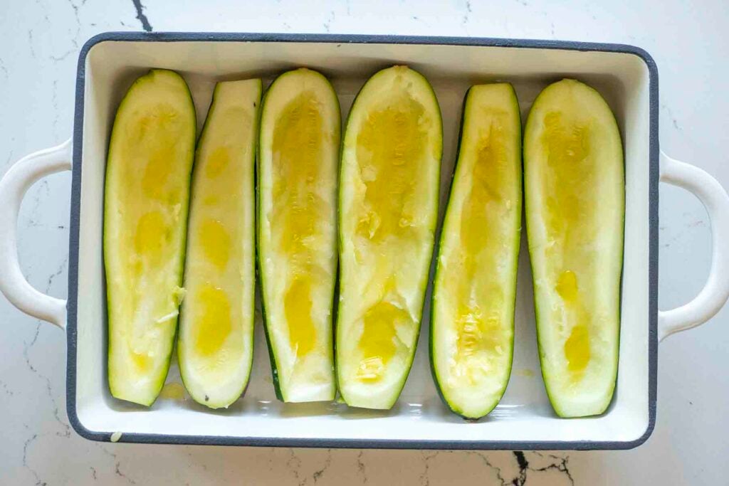 Zucchini halves with the flesh scooped out and drizzled with olive oil in a baking dish.