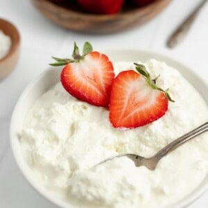 homemade cottage cheese topped with halved strawberries in a white bowl. More strawberries in a bowl in the background.