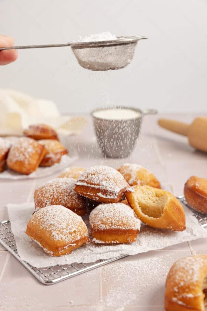 dusting sourdough beignets with powdered sugar on parchment paper.