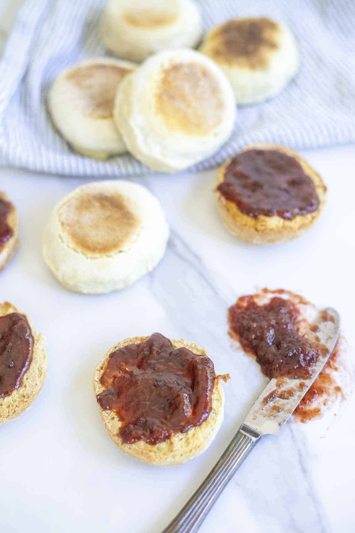 Some sourdough English muffins cut in half with jam and other whole muffins spread on a marble countertop.