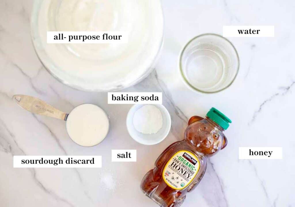 Flour, jar of water, sourdough discard, salt, baking soda, and honey laid out on a countertop.