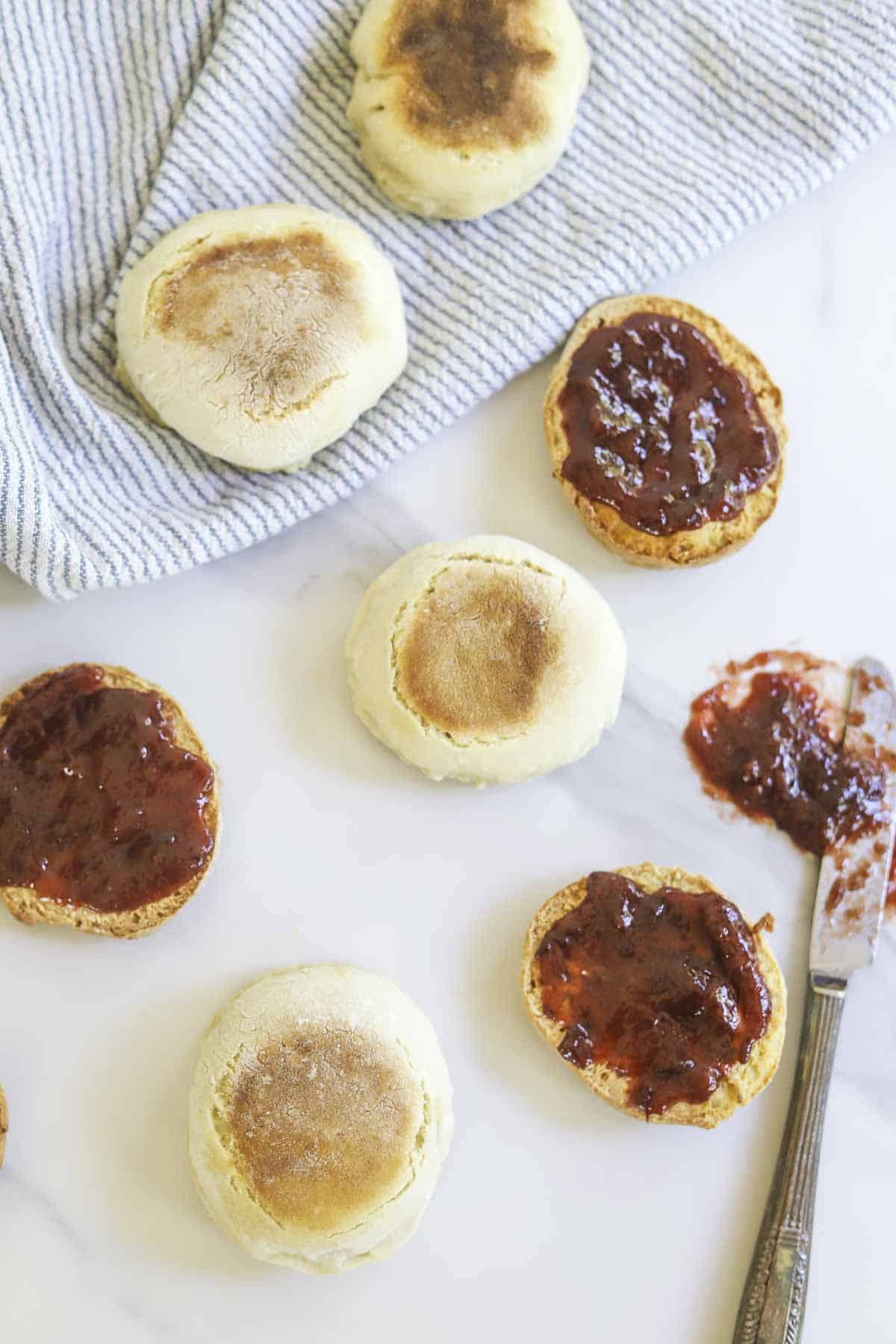 Overhead photo of sourdough English muffins spread out on a countertop. Some of the muffins are cut in half with jam on top.