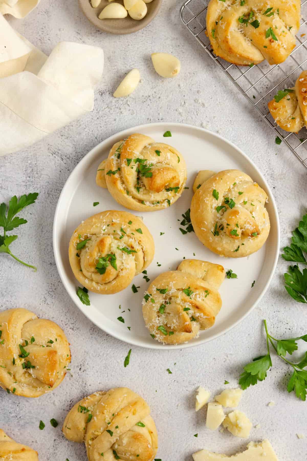 A plate with four sourdough garlic knots topped with parsley. The plate is surrounded with more knots, fresh parsley, garlic, and flour.