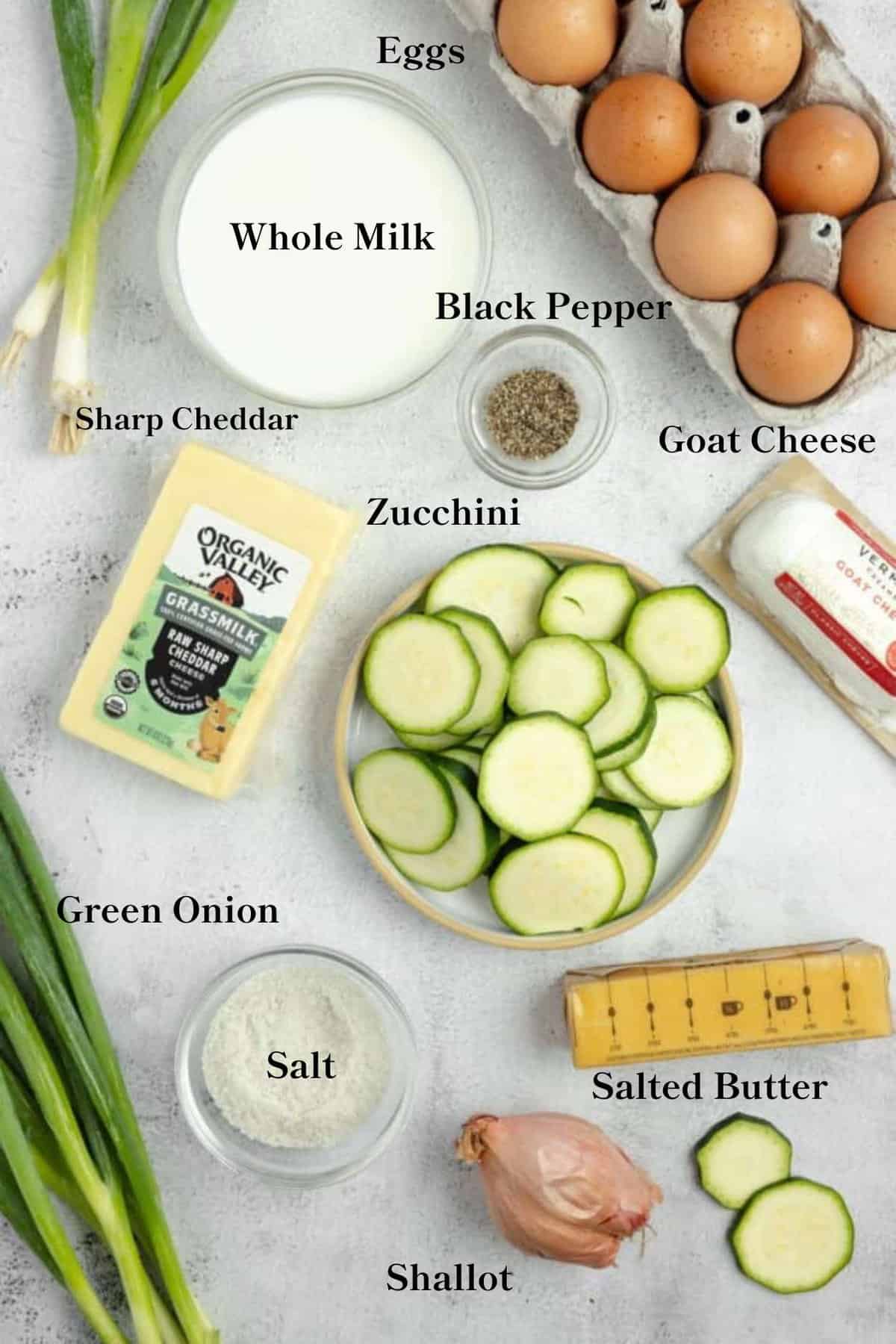 Zucchini frittata ingredients on a countertop. Sliced zucchini, whole milk, eggs, butter, cheese, shallot, salt, green onions, and black pepper spread out on a countertop.