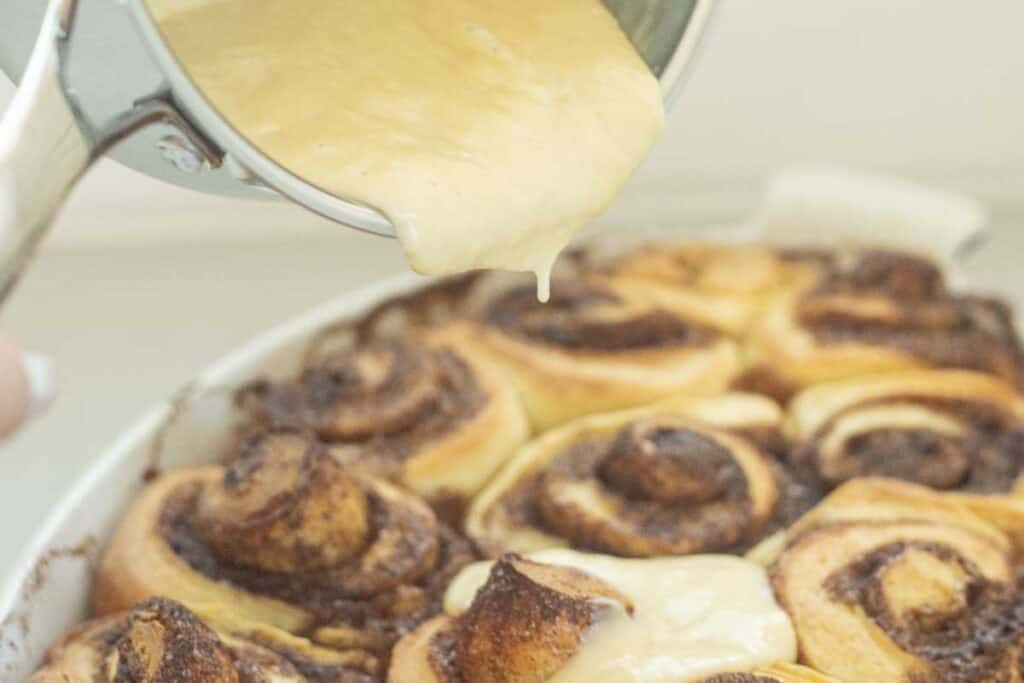 Pouring cream cheese frosting over baked cinnamon rolls.