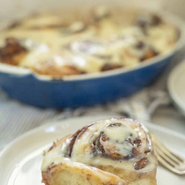 Sourdough brioche cinnamon roll on a white plate with a blue baking pan of more rolls in the background.