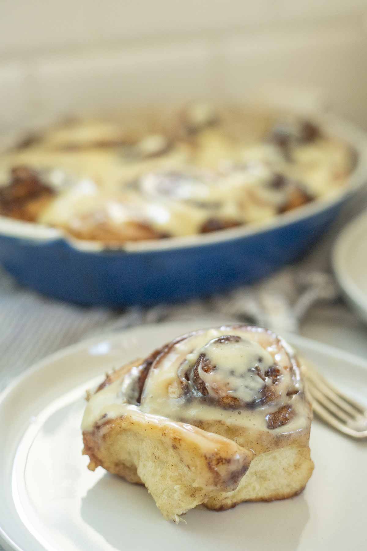 Sourdough brioche cinnamon roll on a white plate with a blue baking pan of more rolls in the background.