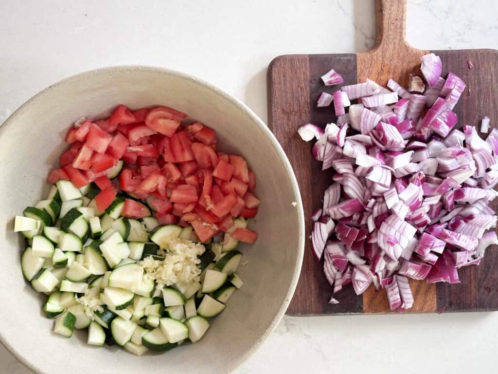 Chopped onion on a cutting board next to a bowl of chopped zucchini and tomato. 