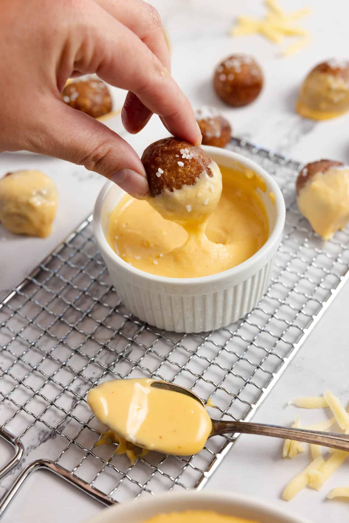 hand dipping pretzel into cheese sauce on a wire rack.