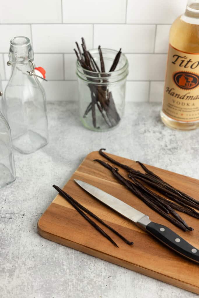 vanilla bean pots cut in half with a knife on a wood cutting board. More vanilla beans in a glass jar, a bottle of vodka and a swing top glass jar in the background.