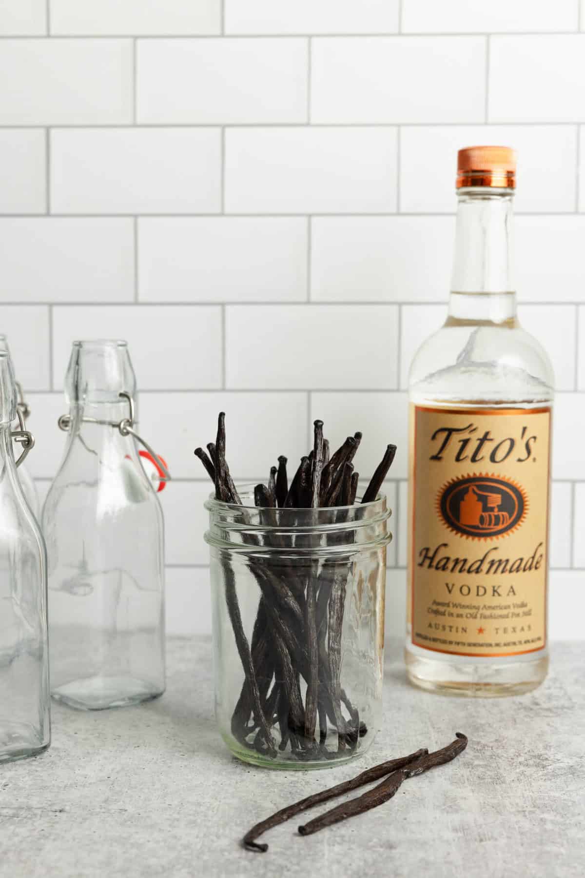 Vanilla beans in a jar, a bottle of vodka, and swing top glass jars on a countertop.