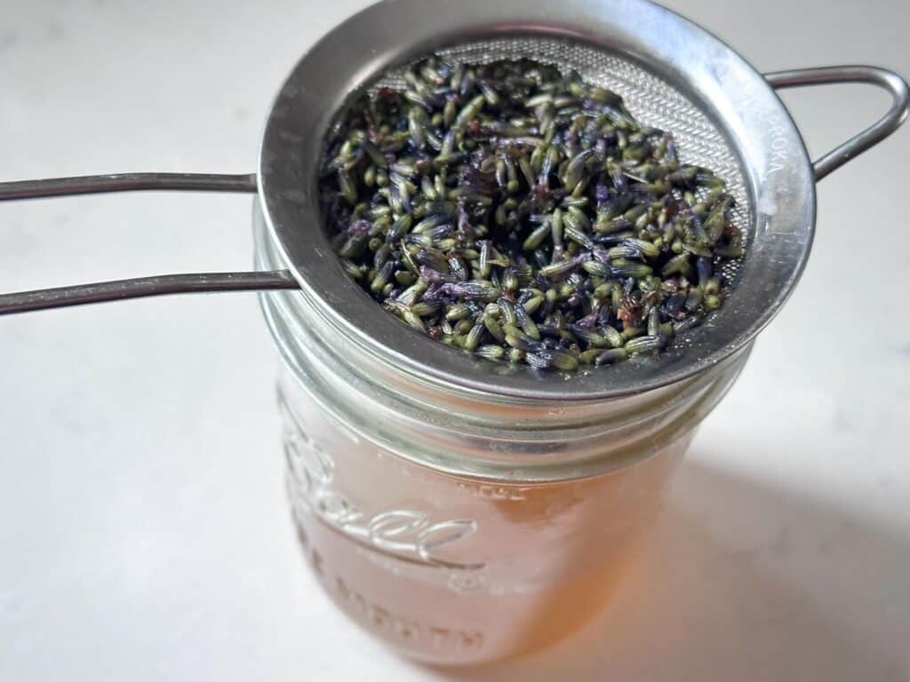 A strainer full of lavender on top of a jar filled with lavender syrup.