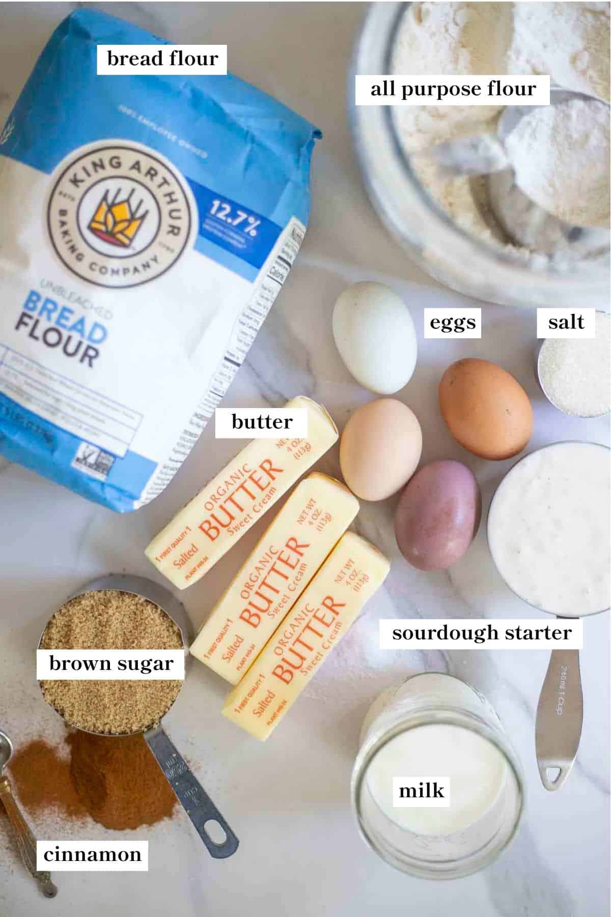 Flour, butter, eggs, milk, salt, brown sugar, cinnamon and more ingredients on a white countertop.