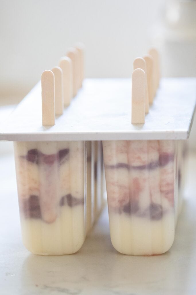 A popsicle mold layered with yogurt and strawberry jam with a lid and popsicle sticks.