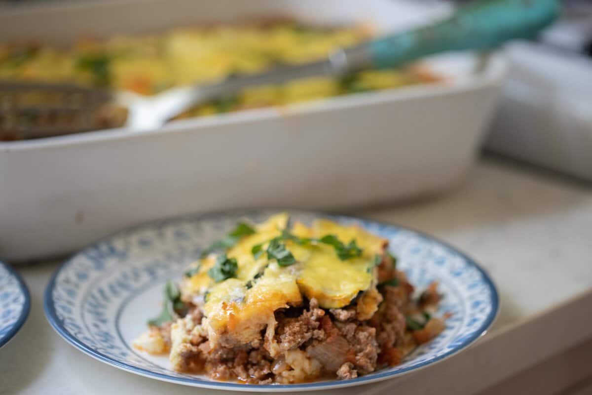 zucchini lasagna on w white plate with a baking dish of more lasagna in the background.