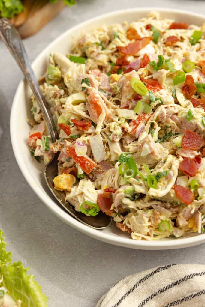 A white serving bowl full of chicken salad with a silver spoon in it.