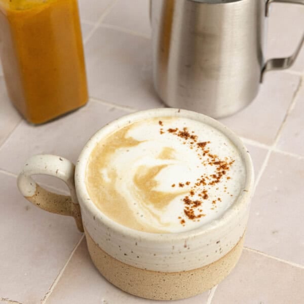 A latte in a white mug next to a silver pitcher filled with steamed milk and a bottle of pumpkin spice sauce.